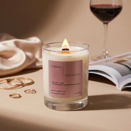 Upsensed Intention candle in a clear glass vessel with a lit wooden wick, surrounded by gold rings, an open magazine, and a glass of red wine on a warm beige surface