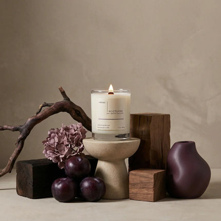 Nocturne candle with plum, florals, and wood accents in an editorial home fragrance still life.