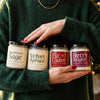 A person in a dark green sweater holding four Upsensed holiday candles in front of softly glowing Christmas lights