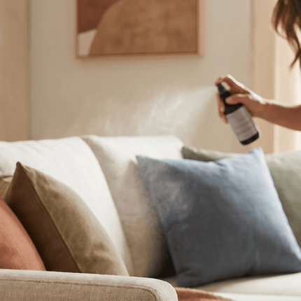 Woman spraying Upsensed luxury home fragrance room spray over a styled linen sofa in a warm, neutral living room.