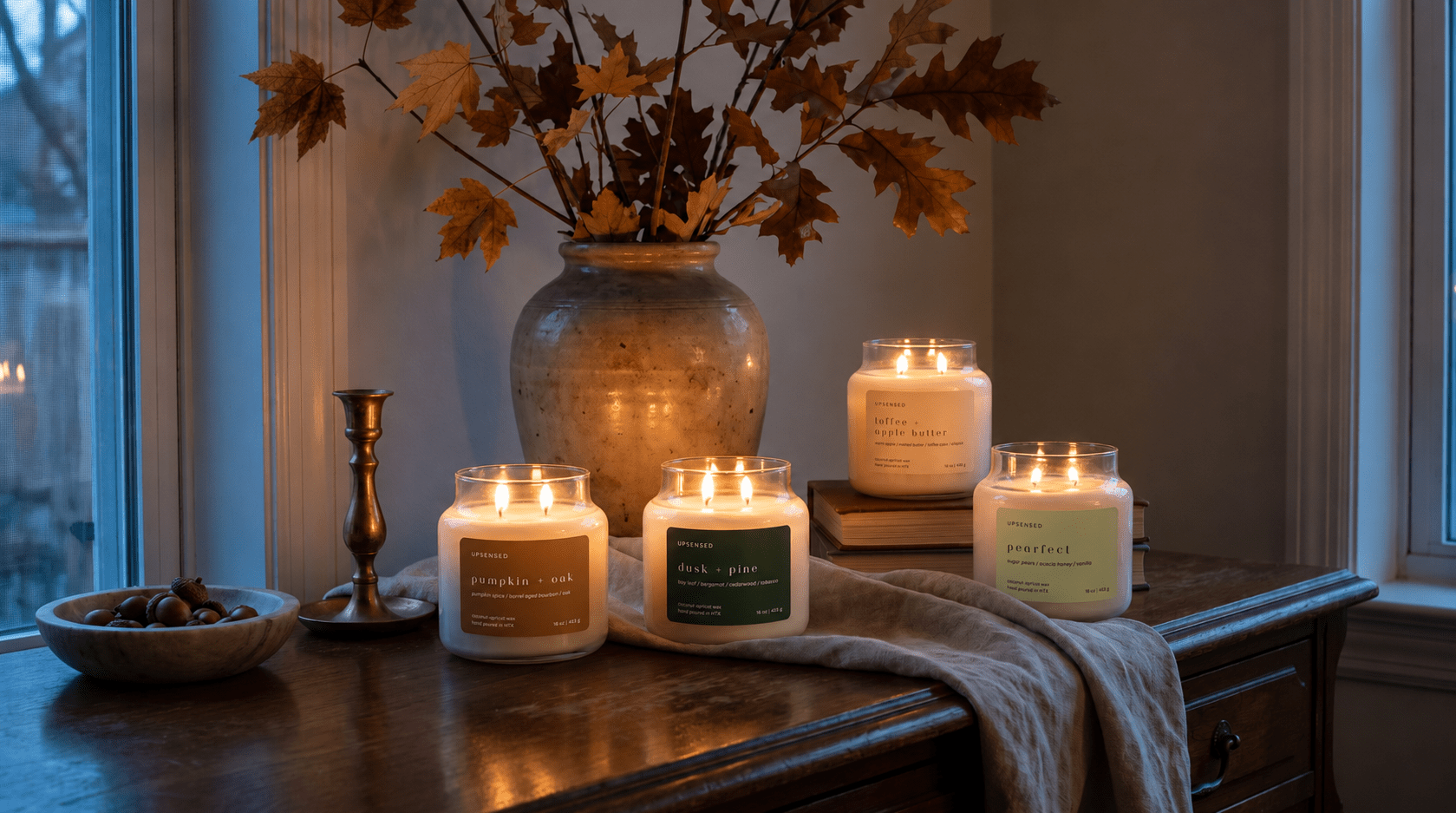Four Upsensed fall candles — Pumpkin + Oak, Pearfect, Dusk + Pine, and Toffee + Apple Butter — lit and grouped on a round wooden riser, surrounded by white pumpkins, scattered fall leaves, and a knit throw.
