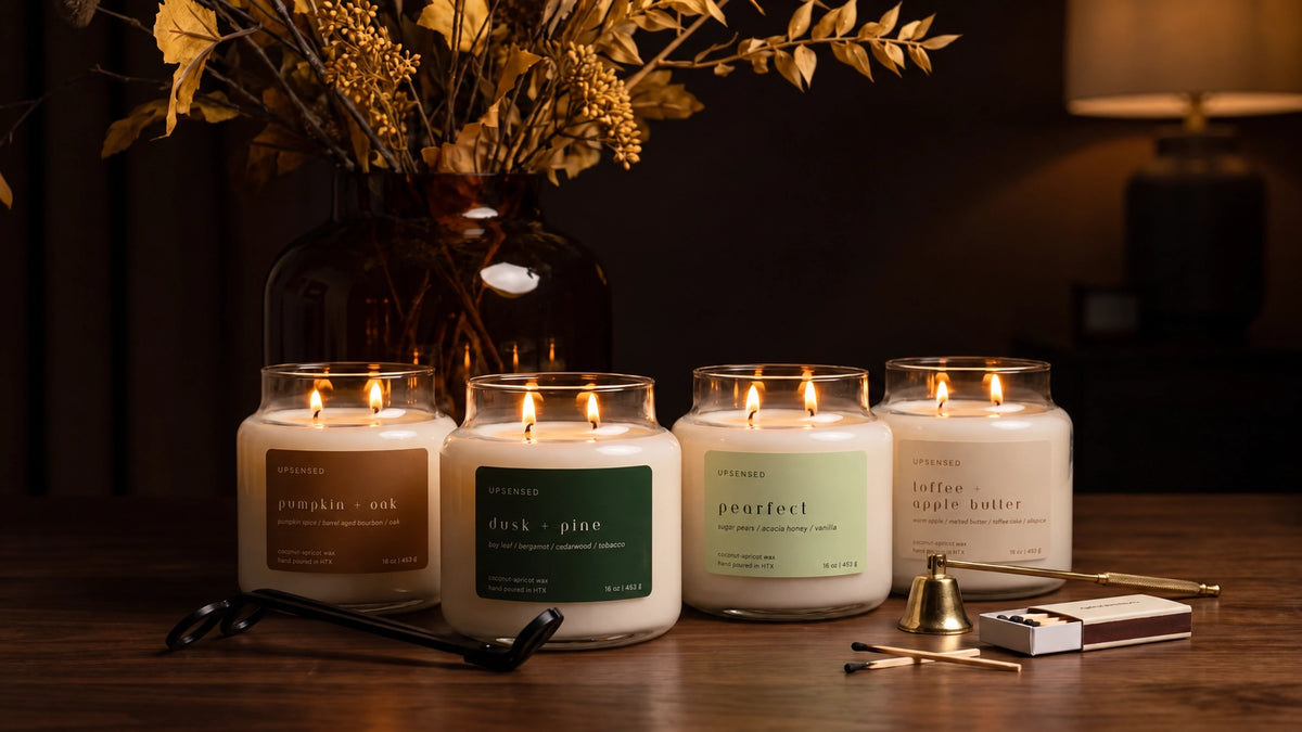 Upsensed fall candle collection featuring Pumpkin + Oak, Dusk + Pine, Pearfect, and Toffee + Apple Butter burning on a dark walnut table with golden autumn branches, matches, wick trimmer, and brass snuffer.