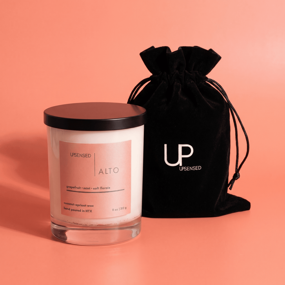 Alto candle in a clear glass jar with a black lid beside a black Upsensed velvet drawstring bag on a coral background.