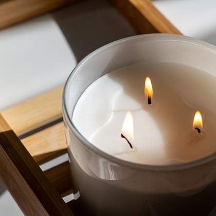 A close-up of a lit three-wick candle in a frosted clear glass jar, resting on a light wood tray.