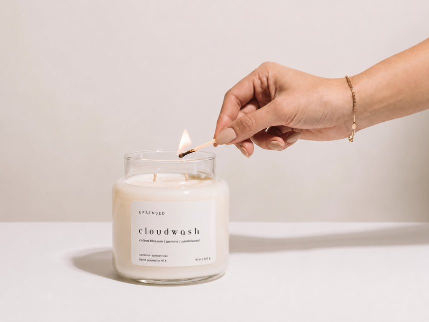 Hand with a gold bracelet lighting an Upsensed Cloudwash candle with a match against a plain background, cotton blossom, jasmine, and sandalwood scent notes visible on the label.