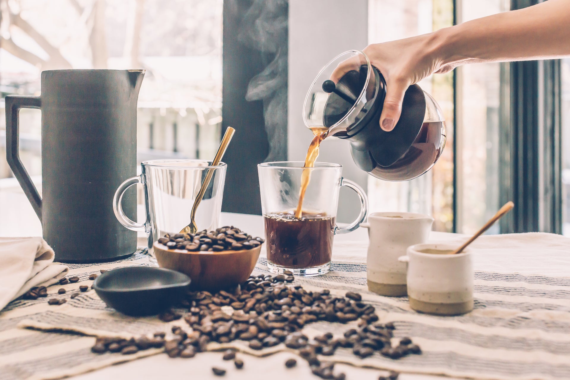 Hand pouring coffee from a French press into a glass mug surrounded by scattered coffee beans and ceramic vessels