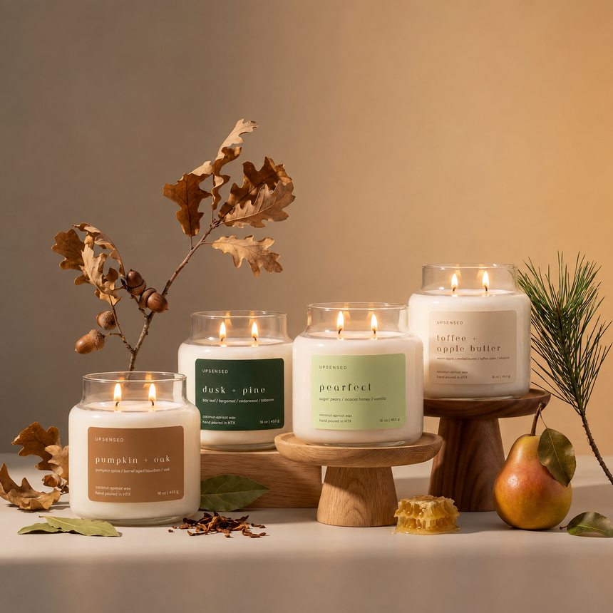 Four lit candles in clear glass jars with white wax and minimalist labels, styled on wooden risers with dried oak leaves, a pine branch, and a pear against a warm beige background.