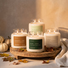 Four Upsensed fall candles — Pumpkin + Oak, Pearfect, Dusk + Pine, and Toffee + Apple Butter — lit and grouped on a round wooden riser, surrounded by white pumpkins, scattered fall leaves, and a knit throw.