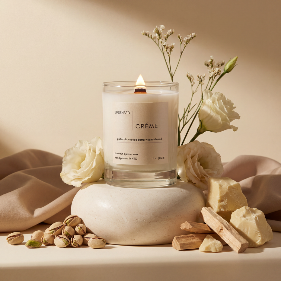 Upsensed Crème 11oz scented candle burning with wood wick surrounded by pistachios cocoa butter sandalwood sticks and white flowers - coconut apricot wax candle hand poured in Houston
