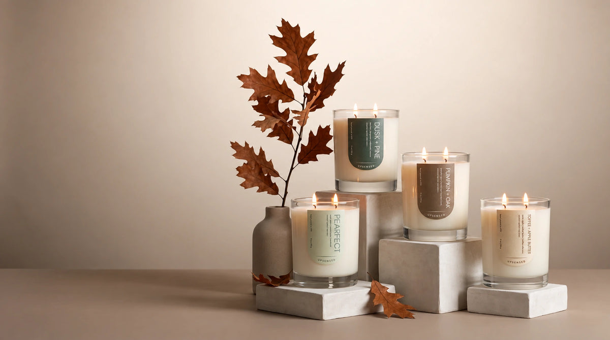 Four lit scented candles in clear glass jars with cream wax, styled on white blocks beside a small vase with brown oak leaves against a warm beige background.