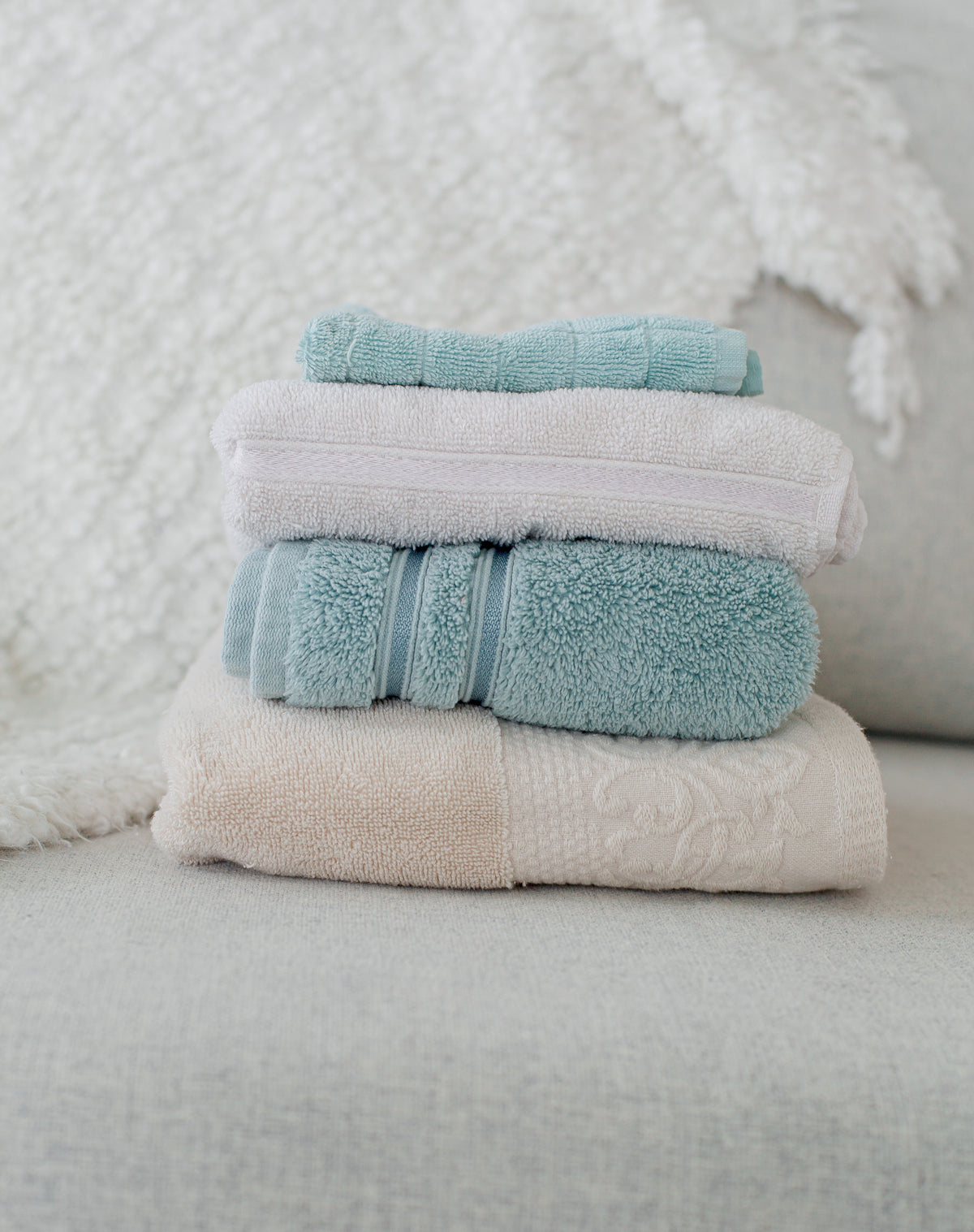Stack of freshly folded sage and blush towels on a cream textured couch — evoking clean, soft home fragrance and everyday luxury ritual