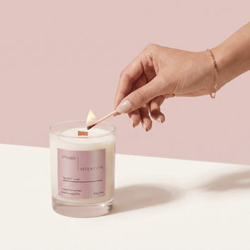 Hand lighting the Intention candle with a match against a soft pink background.