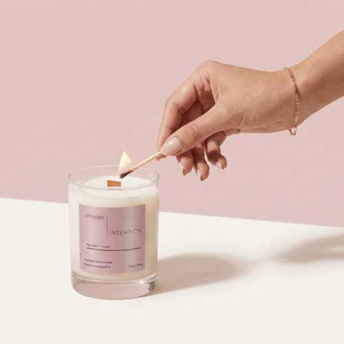 Hand lighting the Intention candle with a match against a soft pink background.