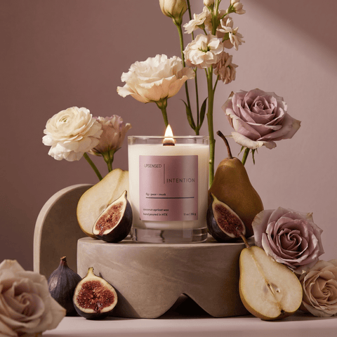 Lit Intention candle styled with fresh figs, sliced pears, soft pink roses, cream flowers, and warm neutral decor.