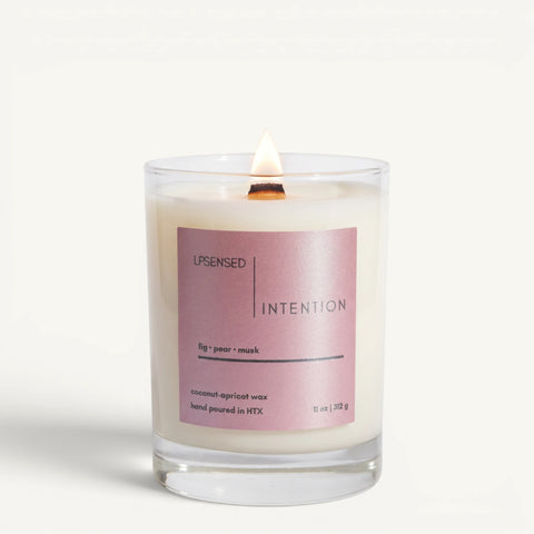 Lit Intention candle in a clear glass jar with a pink label and wooden wick on a clean white background.
