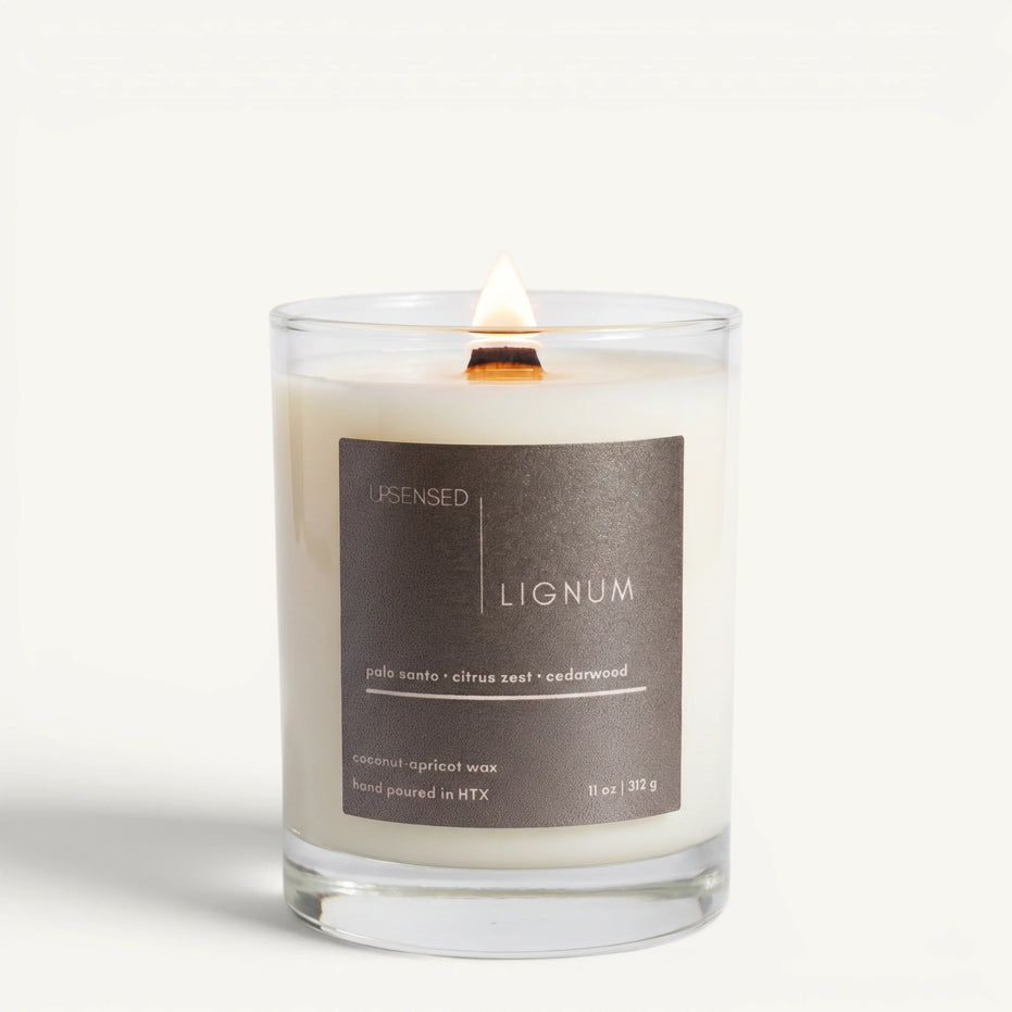 Lit Lignum candle in a clear glass jar with a wooden wick and warm taupe label on a clean white background.