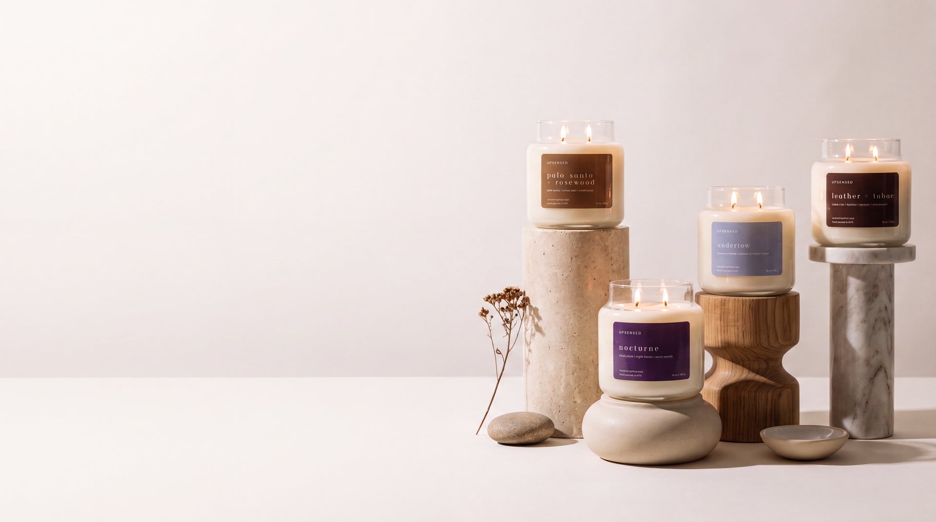 Four lit Upsensed candles — Palo Santo + Rosewood, Nocturne, Undertow, and Leather + Tabac — displayed at staggered heights on stone, wood, and marble pedestals with a dried botanical sprig, against a wide white background with copy space on the left.
