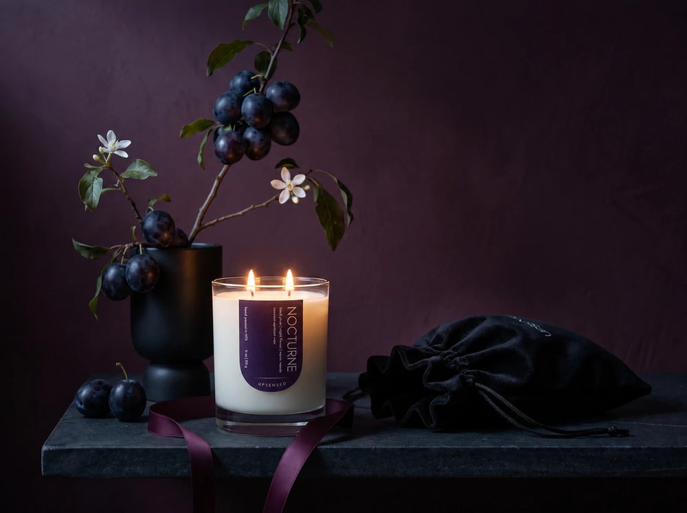Lit Upsensed Nocturne candle on a dark stone ledge with a plum branch, loose plums, plum ribbon, and a black velvet gift pouch