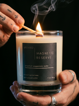 Close-up of a hand lighting the Magnetic Reserve candle with a match, showing the wooden wick, flame, and soft smoke against a dark background.