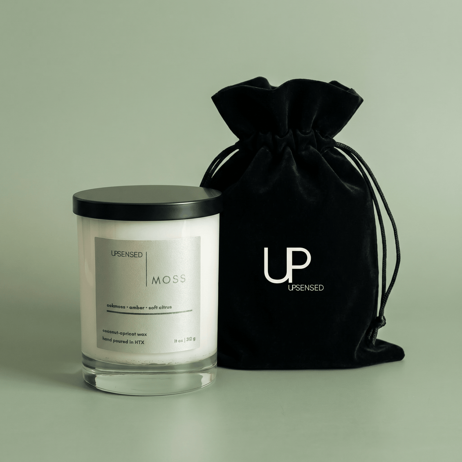 Moss candle in a clear glass jar with a black lid beside a black Upsensed velvet drawstring bag on a soft green background.