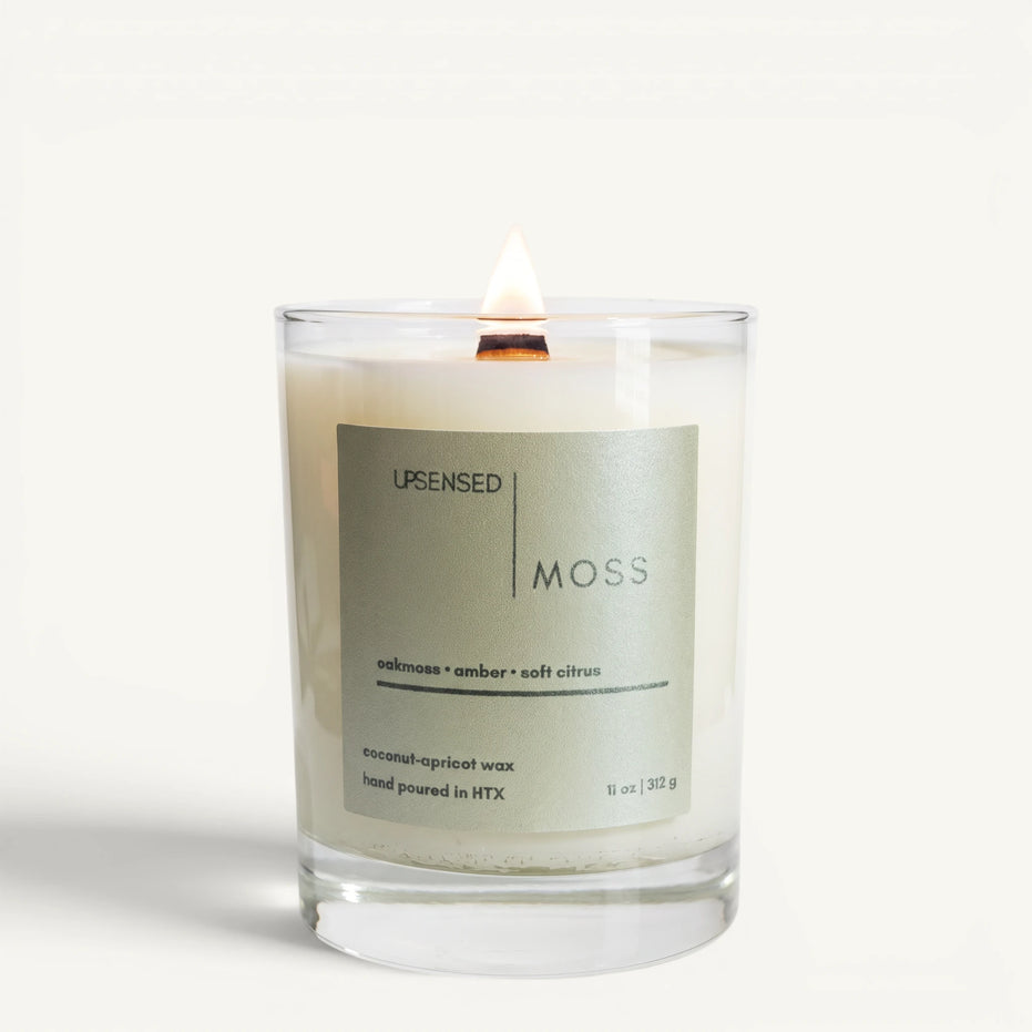 Lit Moss candle in a clear glass jar with a pale green label and wooden wick on a clean white background.