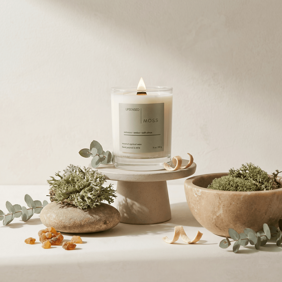 Lit Moss candle styled with oakmoss, eucalyptus stems, amber resin, citrus peel, and warm neutral stone decor.