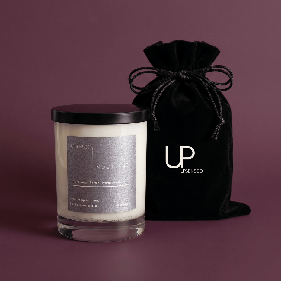 Nocturne candle in a clear glass jar with a black lid beside a black Upsensed velvet drawstring bag on a moody plum background.