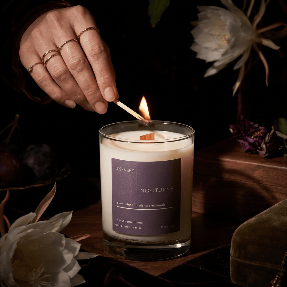 Hand lighting the Nocturne candle with a match in a dark floral setting with plum accents and warm wood details.