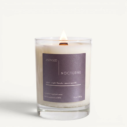 Lit Nocturne candle in a clear glass jar with a deep plum label and wooden wick on a clean white background.