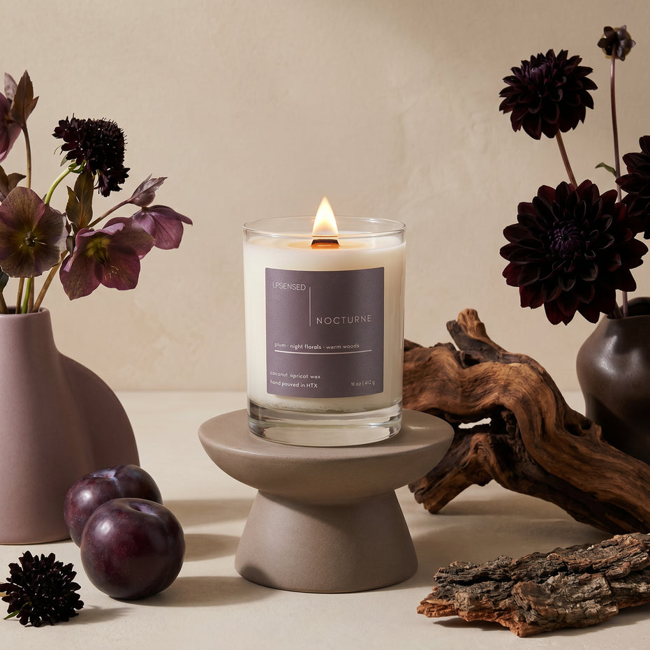 Lit Nocturne candle styled with dark florals, black plums, driftwood, bark, and warm neutral decor.