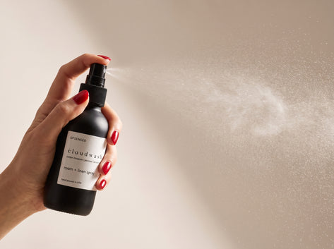 Hand with red manicured nails spraying Cloudwash room and linen spray, fine mist drifting across a warm neutral background.