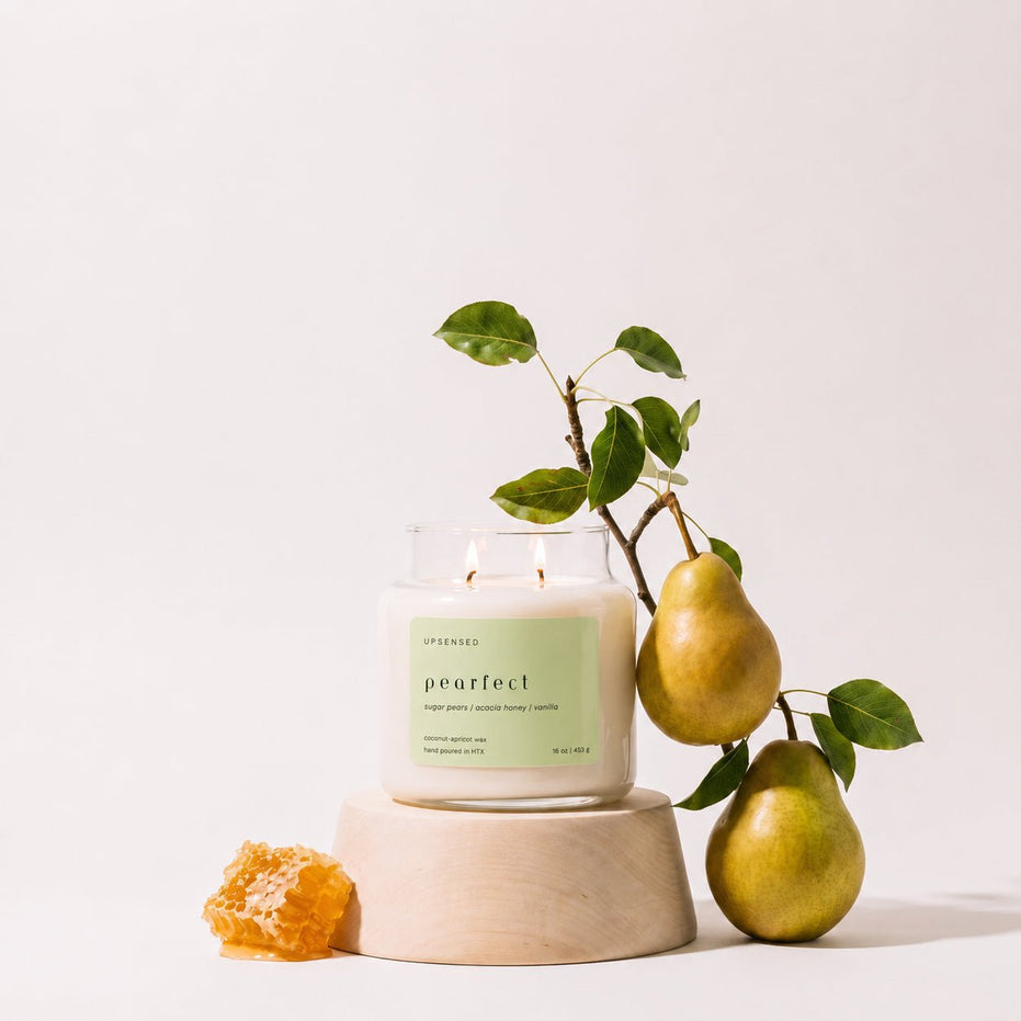Upsensed Pearfect candle burning on a light wood pedestal, styled with a pear branch bearing two pears and a piece of honeycomb.