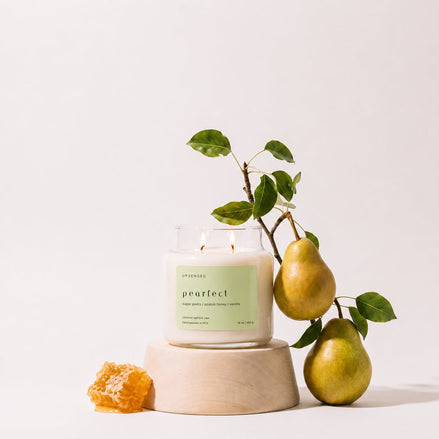 Upsensed Pearfect candle burning on a light wood pedestal, styled with a pear branch bearing two pears and a piece of honeycomb.