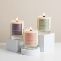 Three lit Upsensed coconut-apricot wax candles displayed on white risers, featuring Alto, Moss, and Nocturne in clear glass jars.