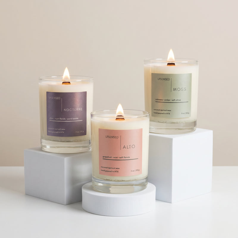Three lit Upsensed coconut-apricot wax candles displayed on white risers, featuring Alto, Moss, and Nocturne in clear glass jars.