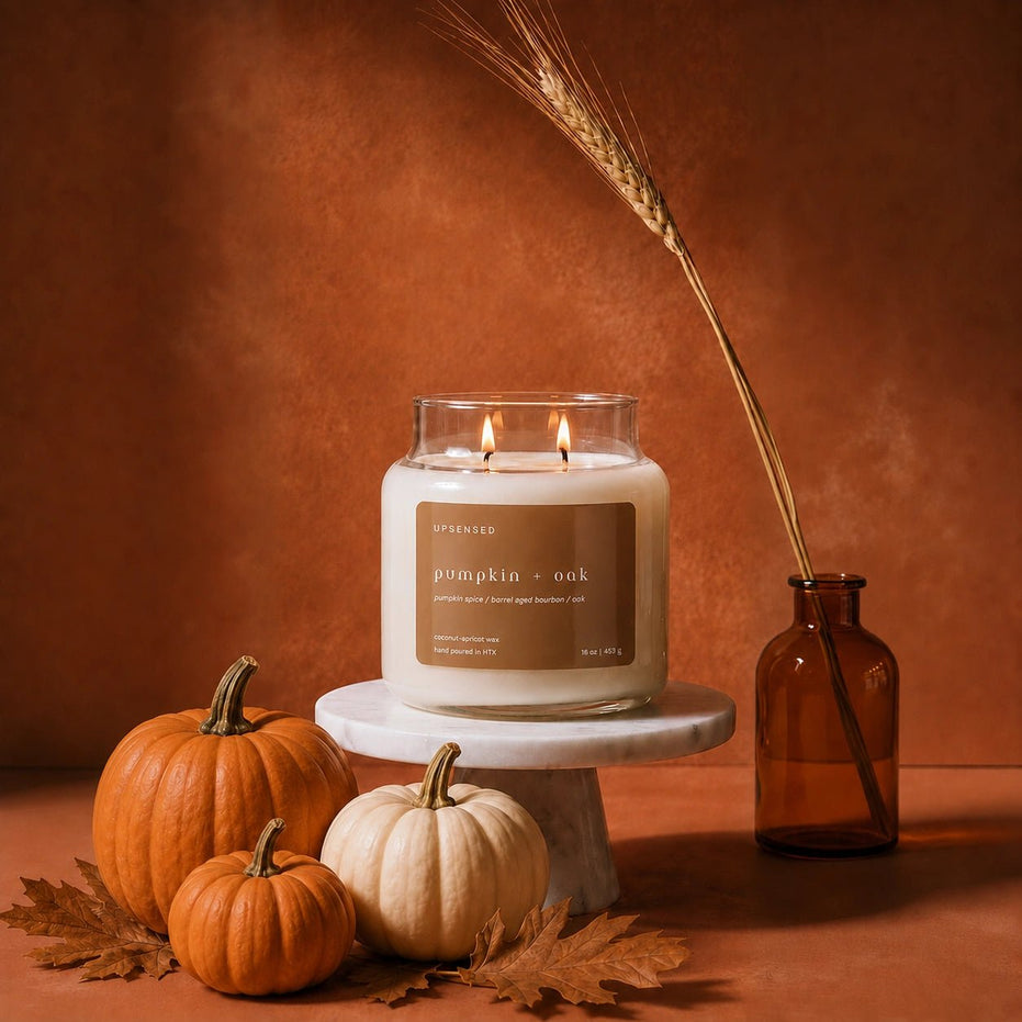 A lit candle in a clear glass jar sits on a white pedestal, surrounded by small pumpkins, autumn leaves, and a tall amber bottle with a wheat stem, against a warm brown textured background. The candle label reads 'UPSENSED' and 'pumpkin + oak'.