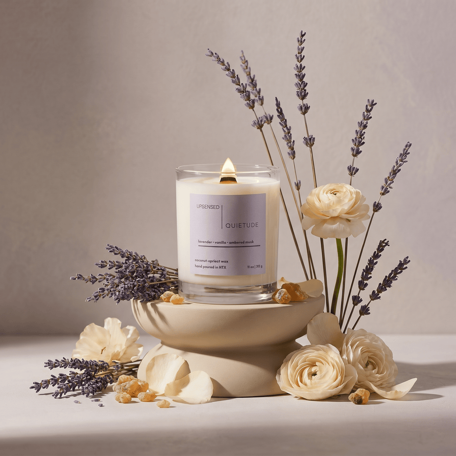 Lit Quietude candle styled with lavender stems, cream flowers, petals, and ambered resin in a soft floral still life.