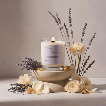 Lit Quietude candle styled with lavender stems, cream flowers, petals, and ambered resin in a soft floral still life.
