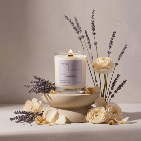 Lit Quietude candle styled with lavender stems, cream flowers, petals, and ambered resin in a soft floral still life.