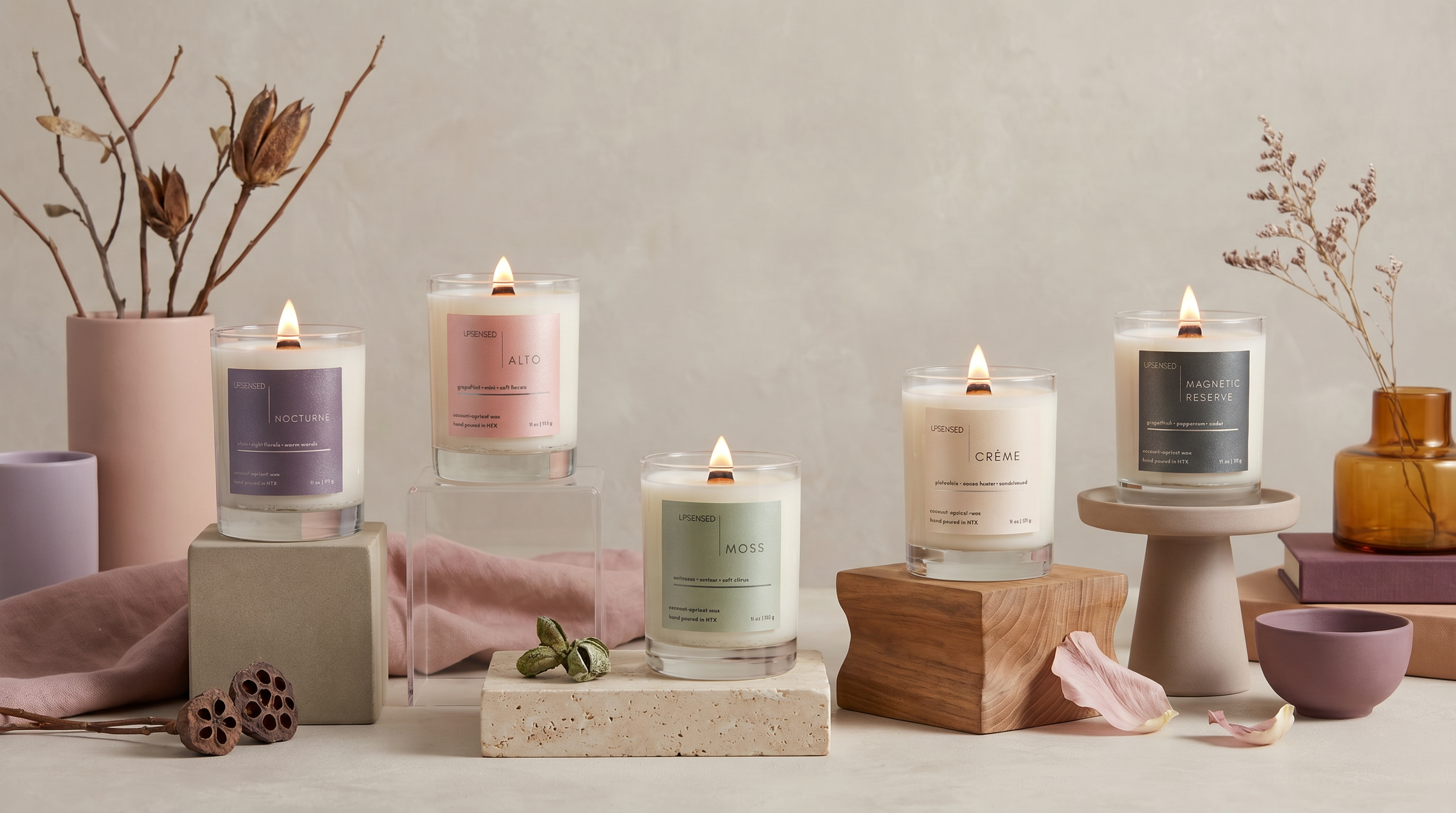 Five lit Upsensed candles arranged on sculptural risers with soft neutral styling, dried botanicals, petals, and warm-toned decor.