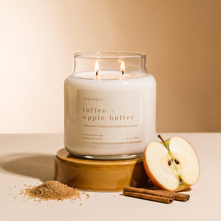 Upsensed Toffee + Apple Butter candle lit next to a halved apple, cinnamon sticks, and a scattering of brown sugar against a warm tan background.