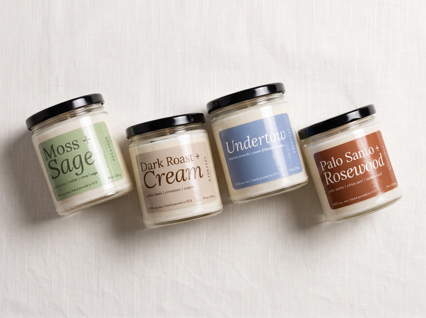 Four glass jar candles with black lids are arranged diagonally on a light textured fabric background. The labels read Moss + Sage, Dark Roast + Cream, Untitled, and Palo Santo + Rosewood.