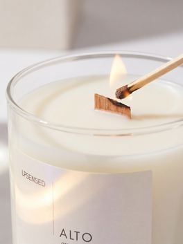 Close-up of an Upsensed Alto wooden wick candle being lit with a match, showing creamy wax, a clear glass vessel, and a glowing pearlescent label.