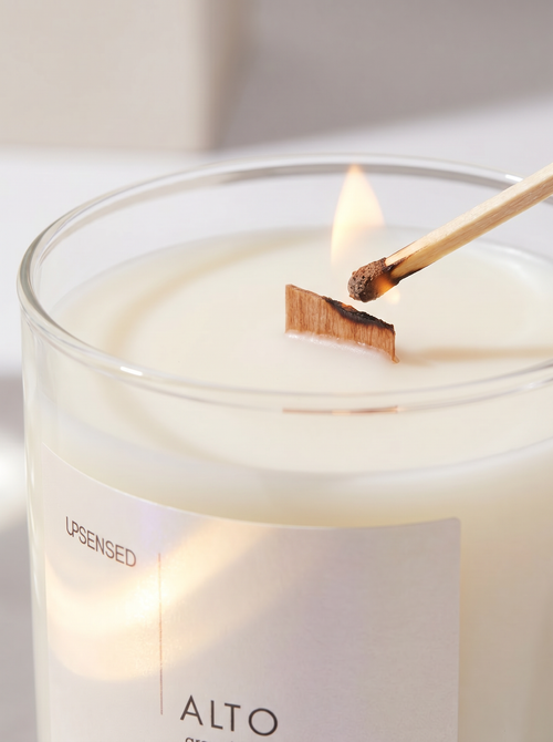 Close-up of an Upsensed Alto wooden wick candle being lit with a match, showing creamy wax, a clear glass vessel, and a glowing pearlescent label.