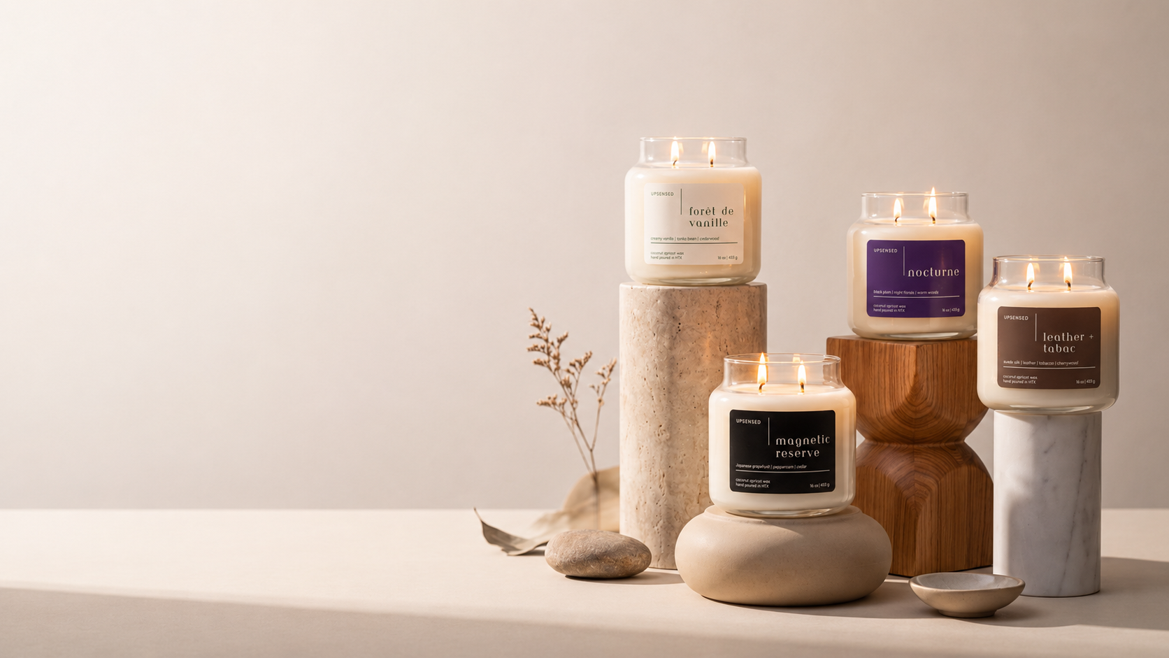 Four lit Upsensed candles — Forêt de Vanille, Nocturne, Magnetic Reserve, and Leather + Tabac — displayed at staggered heights on stone, wood, and marble pedestals with dried botanicals.