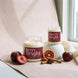 Two lit candles labeled Berry Bright are styled on a soft beige surface near a window, with dark berries, a sliced berry, cinnamon sticks, and star anise around them.