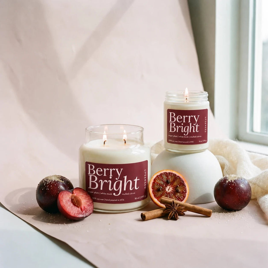 Two lit candles labeled Berry Bright are styled on a soft beige surface near a window, with dark berries, a sliced berry, cinnamon sticks, and star anise around them.