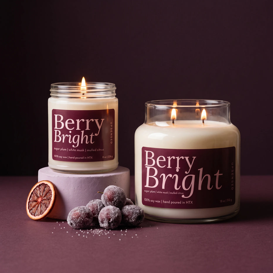 Two lit scented candles labeled Berry Bright are displayed on a dark purple surface against a matching dark background. One candle is in a clear glass jar resting on a pale lavender pedestal, and the other is in a larger clear glass jar.