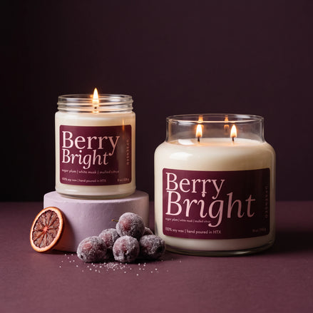 Two lit scented candles labeled Berry Bright are displayed on a dark purple surface against a matching dark background. One candle is in a clear glass jar resting on a pale lavender pedestal, and the other is in a larger clear glass jar.