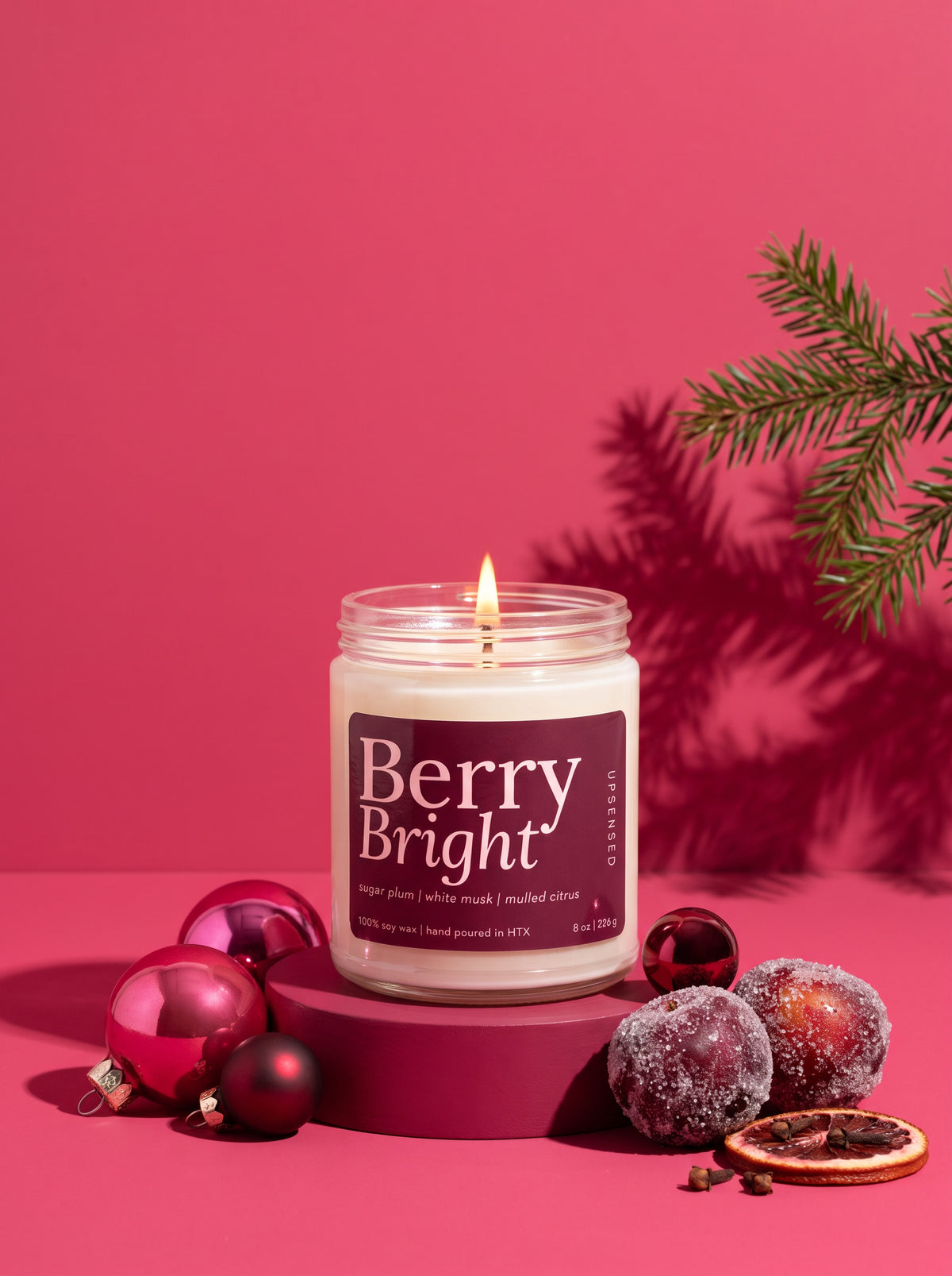 A lit candle in a clear glass jar labeled 'Berry Bright' sits on a magenta pedestal against a pink background, styled with red ornaments, frosted berries, a dried orange slice, and pine branches.